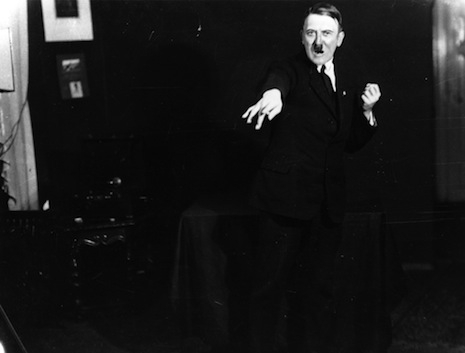 black and white photograph of hitler facing the camera with his left hand clenched in a fist near his left shoulder and his right hand gesturing away from his body with one finger pointed out and the others hanging limply.